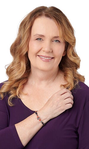 A smiling woman with wavy strawberry-blonde hair wears a beaded bracelet featuring black, red, and silver Halloween charms.