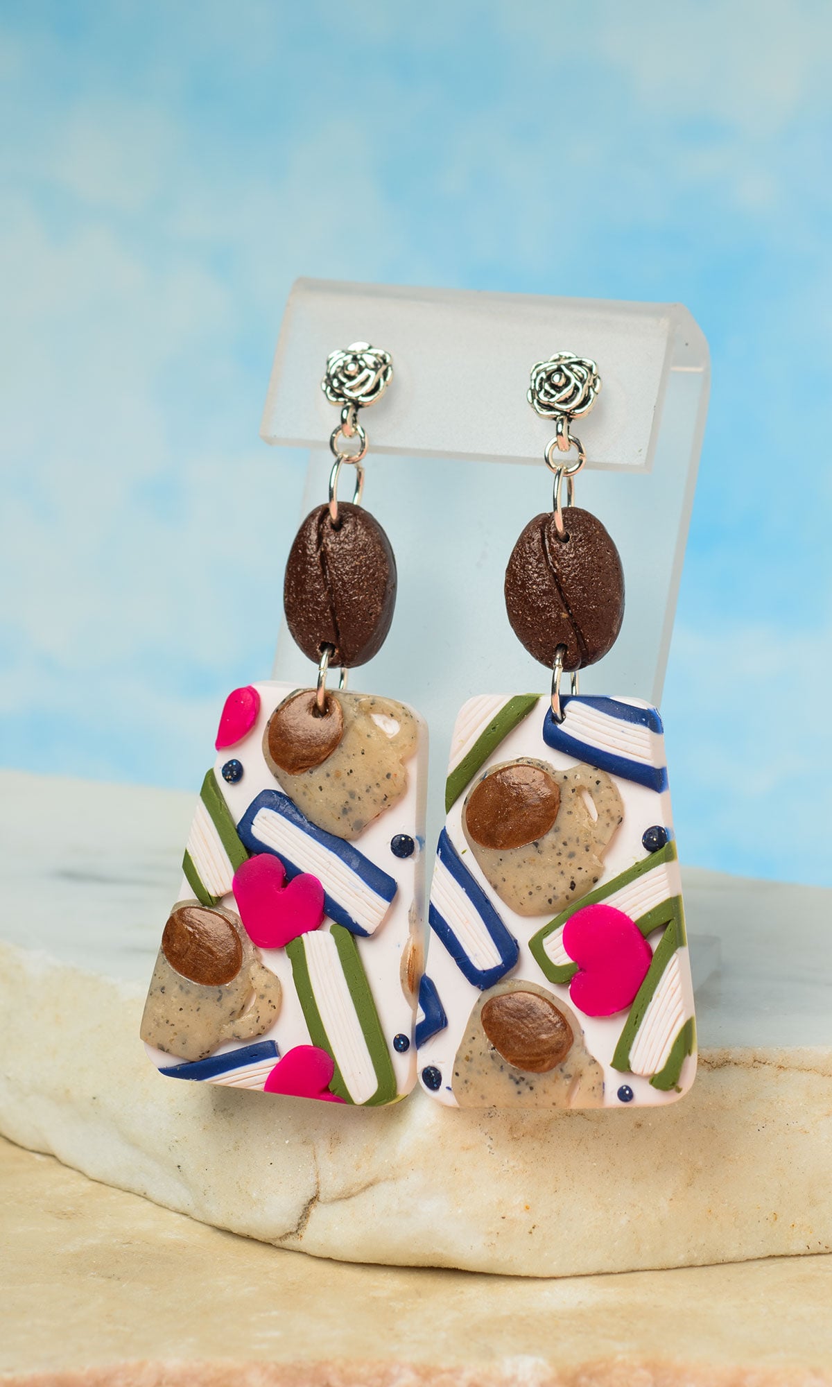 A pair of dangling polymer clay earrings featuring simulated brown coffee beans, books, mugs, and pink hearts.