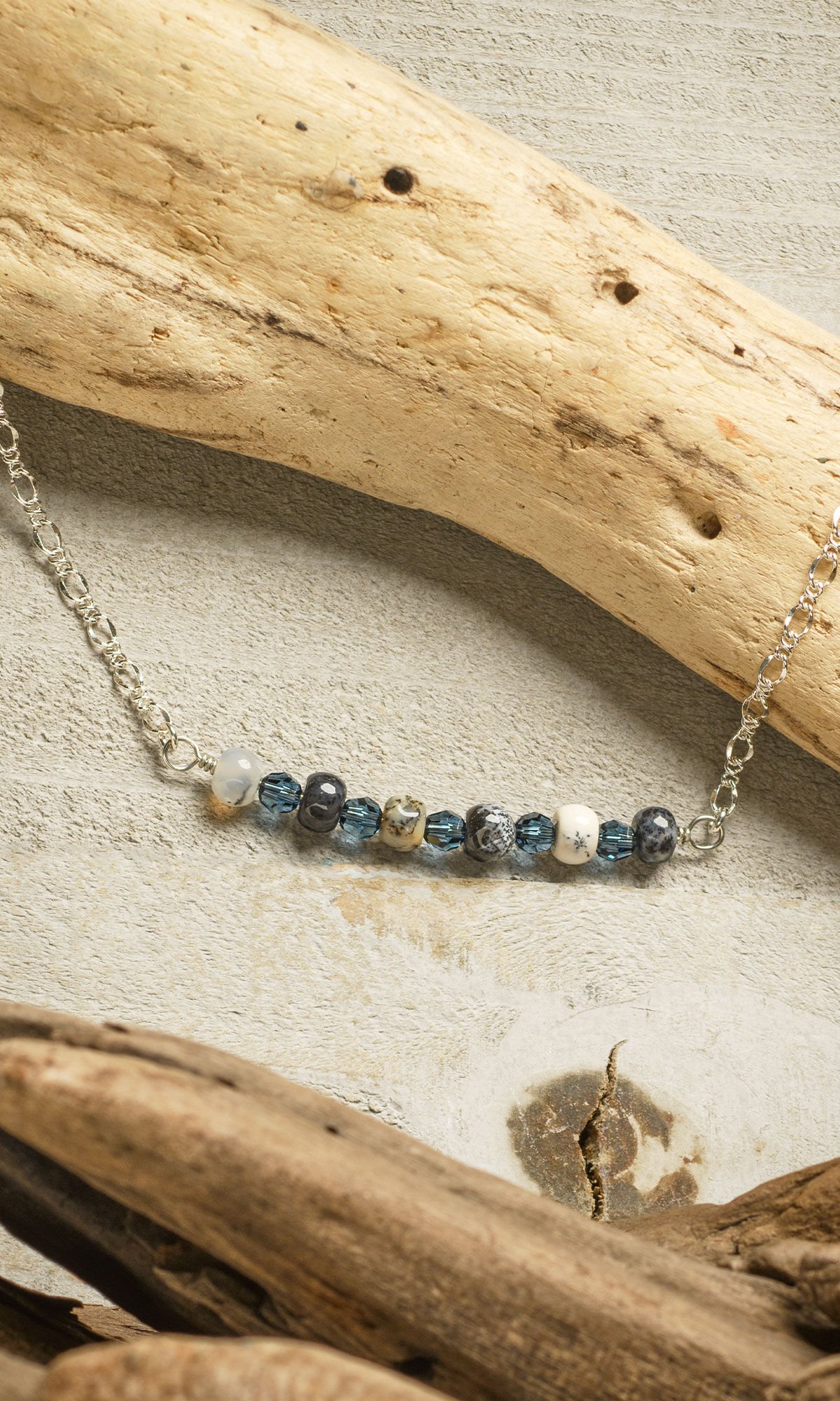 Design Idea R G 2 N Silver chain necklace with a flat horizontal length of blue and multicolored beads at the front lying on a piece of driftwood with a tan background