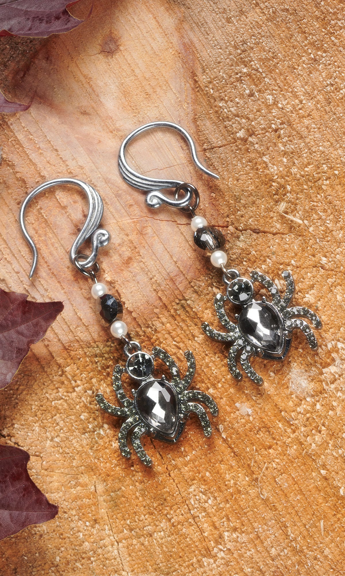 Silver-toned drop earrings shaped like spiders, embellished with grey crystals and small pearls, resting on a rustic wood surface.
