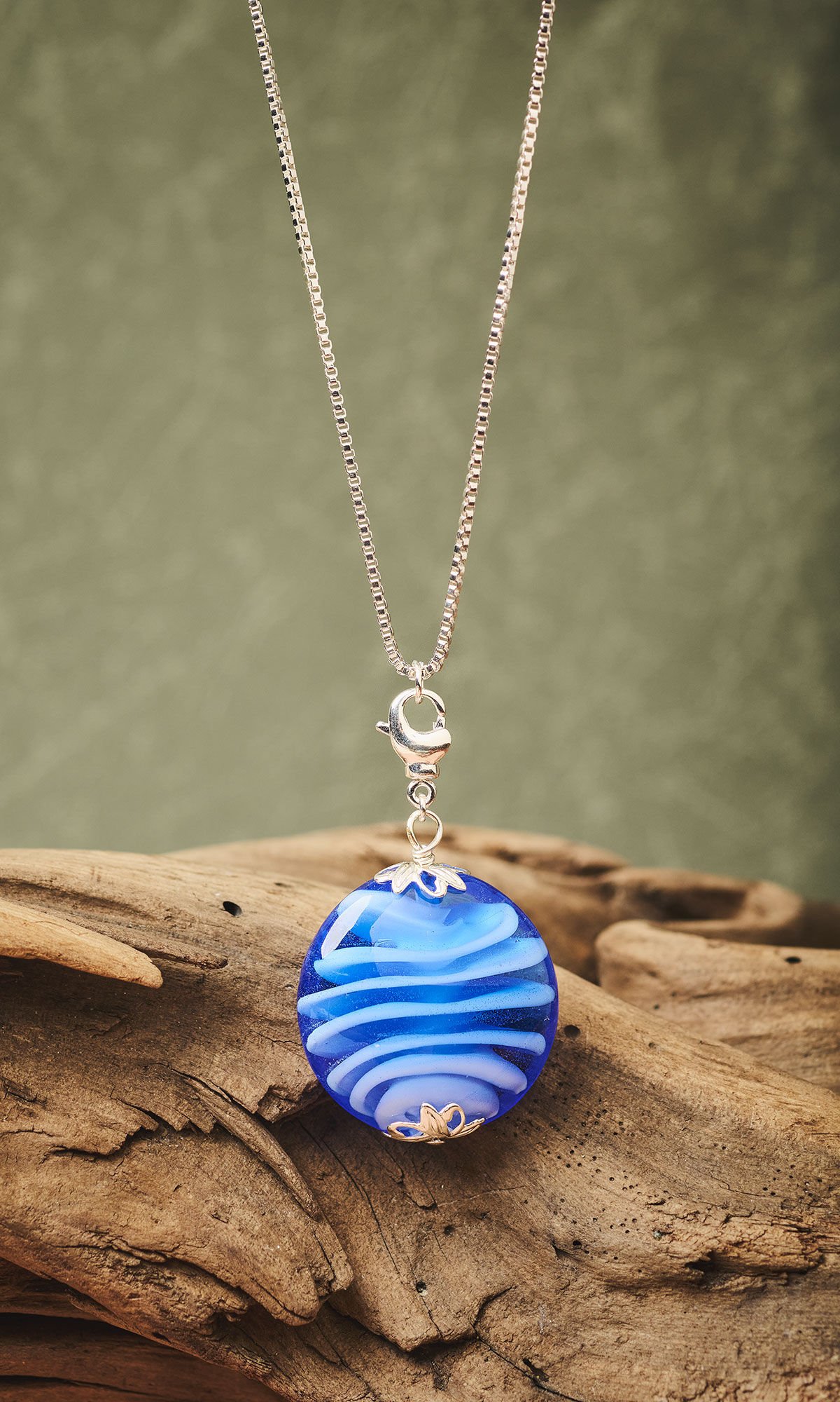 Chain necklace with blue lampworked glass pendant hanging in front of driftwood